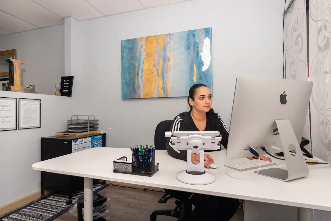 A woman sits at a desk working on an Apple desktop computer in a modern office, with pens, papers, and wall art in the background.