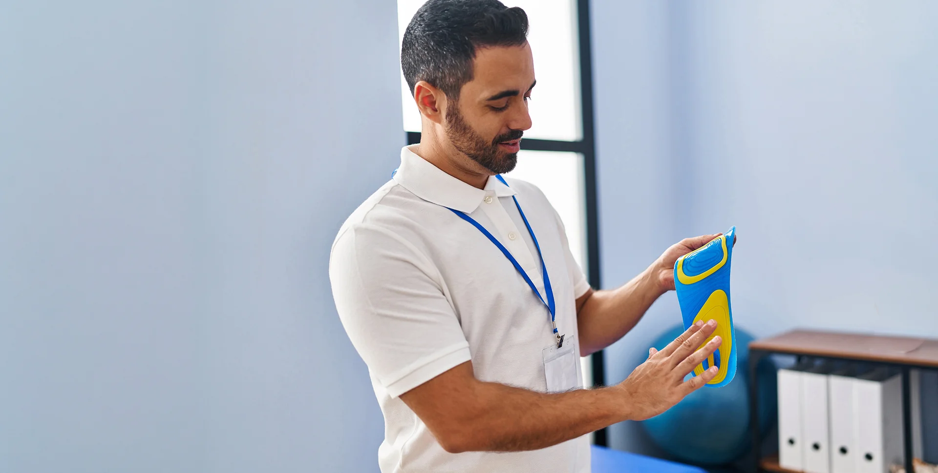 A male healthcare professional wearing a white polo shirt and lanyard inspecting a blue and yellow orthopedic shoe insole.